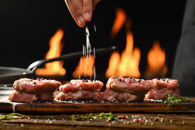 生のテンダーロインステーキ肉牛肉に塩と調味料を手で振りかけ、背景の炎で調理するために準備された木製のテーブルの木製まな板に