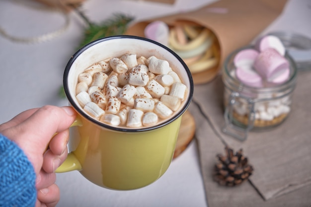 A hand in a sweater holds a yellow cup with cocao decorated with marshmallows, a stand made of saw cut wood and marshmallows on the background