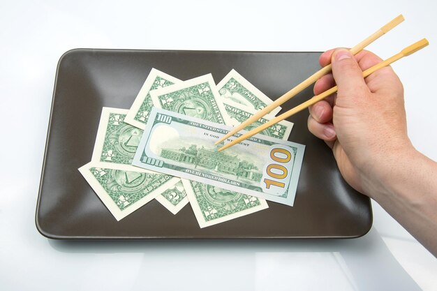 Hand takes dollars with bamboo chopsticks from a ceramic plate financial industry in food cash spending concept