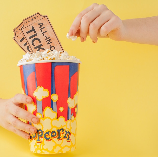 Hand takes a movie tickets and popcorn from a paper cup on a yellow background. Woman eats popcorn. Cinema Concept. Flat lay.