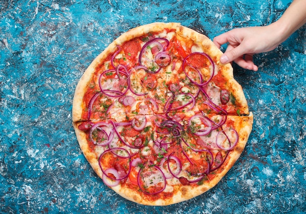 The hand takes a piece of homemade pizza on blue concrete. Top view