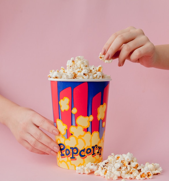 La mano prende un popcorn da un bicchiere di carta su uno sfondo rosa. la donna mangia popcorn. concetto di cinema. disteso. copyspace