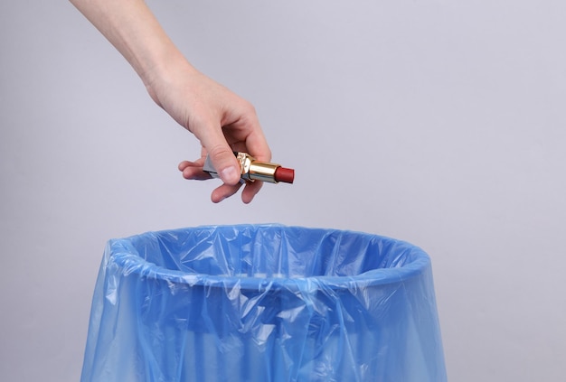 Hand throws lipstick into trash bin with package on gray background