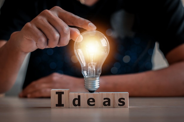 Hand touching on light bulb on wood block with Word Ideas, new idea concept with innovation and inspiration, innovative technology in science and communication concept.