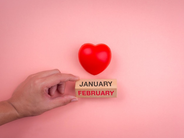 Hand turning wooden cube block for change the month from january to february and red heart ball on pink pastel background. concept of valentine's day, love, minimal style