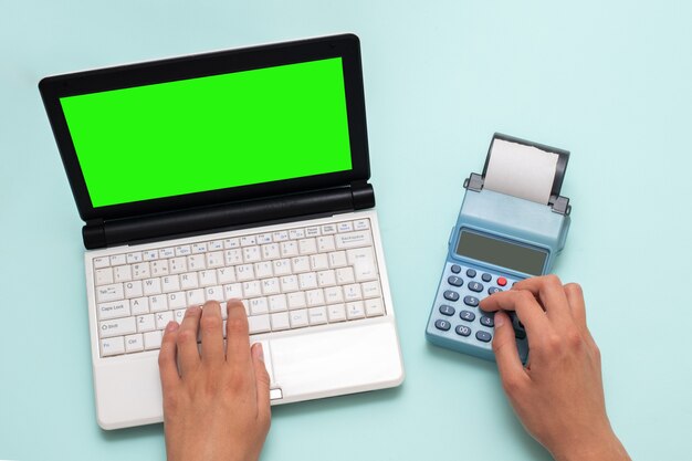 Hand typing on a laptop with a green screen and pressing buttons on a cash register on a blue background, copy space. The cashier selects the item and enters the amount