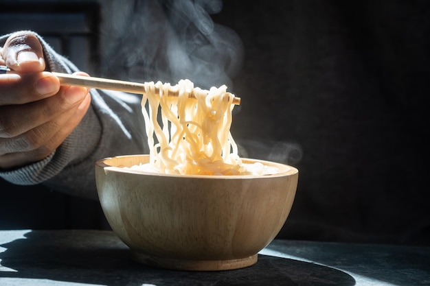 손은 젓가락을 사용하여 나무 배경에 있는 그릇에 증기와 연기가 있는 맛있는 국수를 집어들며 테이블 정크 푸드 개념에 대한 아시아 식사를 선택적으로 집중합니다.