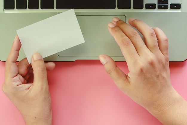 Hand using computer laptop and holding blank business cards on pink background