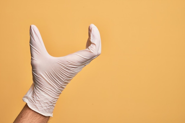 Hand van blanke jonge man met medische handschoen over geïsoleerde gele achtergrond plukken en nemen onzichtbaar ding houden object met vingers tonen ruimte