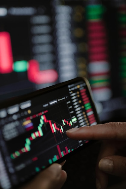 Hand van een jong zakenvrouw die op kantoorcomputer en mobiele telefoon naar handelsgrafieken kijkt