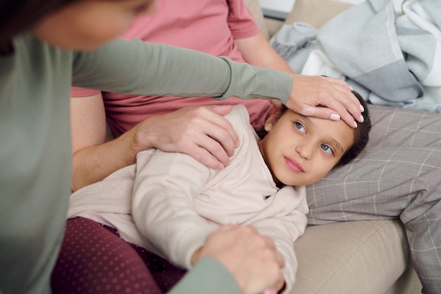 Hand van een jonge, voorzichtige vrouw die voor haar zieke schattige dochter zorgt die op de bank op vaderknieën ligt terwijl ze haar voorhoofd thuis aanraakt