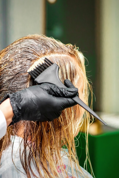 Hand van een kapper in zwarte handschoenen die kleurstof aanbrengt op het vrouwelijke haar in een schoonheidssalon