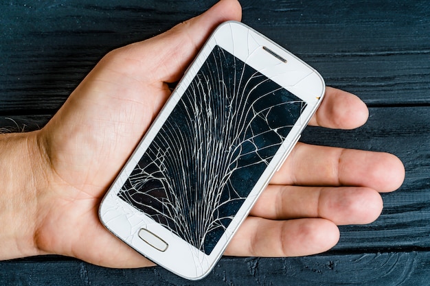 Hand van een mannetje dat witte smartphone met het beschadigde glasscherm binnen houdt.