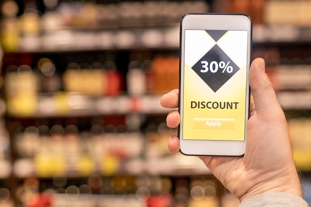 Hand van hedendaagse klant die mobiele app toont die u een goede korting geeft voor het aantal voedselproducten in de supermarkt