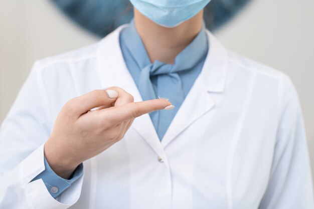 Hand van jonge hedendaagse vrouwelijke oogarts in whitecoat en beschermend masker met contactlens op wijsvinger voor camera