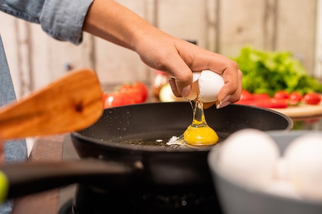 Hand van jonge vrouw die vers ei op koekenpan breekt die zich op elektrisch fornuis bevindt tijdens het koken van ontbijt in de ochtend