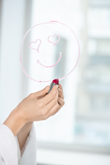 Hand van meisje met karmozijnrode lippenstift tekening gezicht met glimlach en hartvormige ogen op spiegel als liefdesbericht
