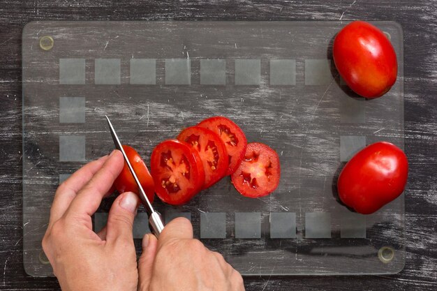 Foto hand van vrouw die verse tomaten met mes snijdt op het snijbord van glas op zwarte achtergrond