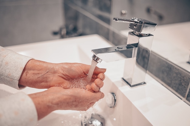 Ручная стирка пены жидкое мыло втирание запястий руки мытье шаг старшая женщина полоскание в воде в ванной кран раковина.