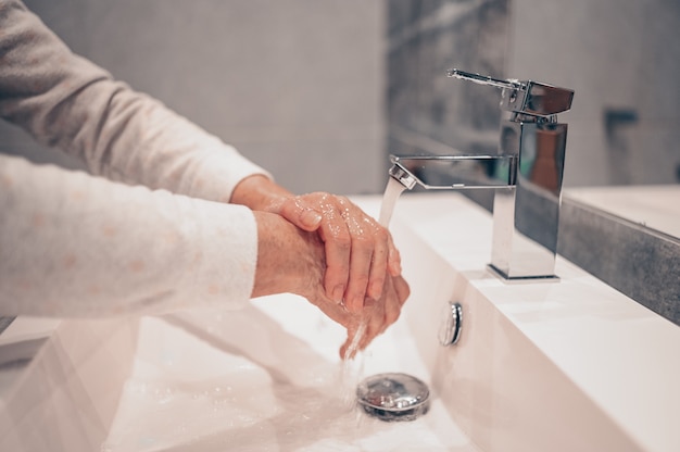 Hand washing lather liquid soap rubbing wrists handwash step senior woman rinsing in water at bathroom faucet sink.