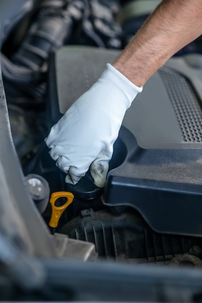 Рука в белой защитной перчатке выполняет скручивающее действие под открытым капотом автомобиля