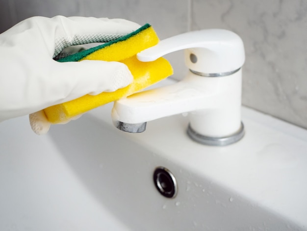 A hand in a white rubber glove with a sponge washes a white faucet in the bathroom