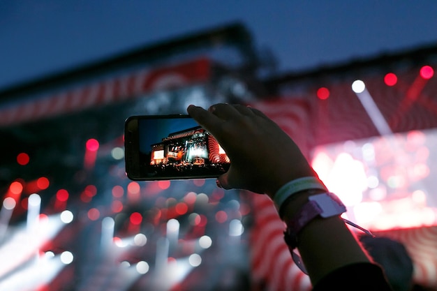 Рука со смартфоном записывает фестиваль живой музыки и фотографирует концертную сцену