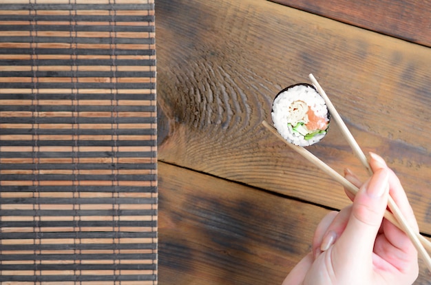 A hand with chopsticks holds a sushi roll on a bamboo straw