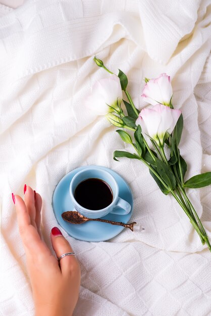 Photo hand with cup of coffee with chocolate, flowers eustoma on blanket in bed.