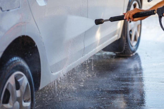 Hand with high pressure washer washing white car at public\
selfservice car washing station closeup with selective focus