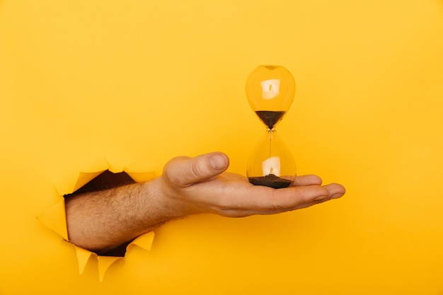 Photo hand with hourglass through a hole of yellow background