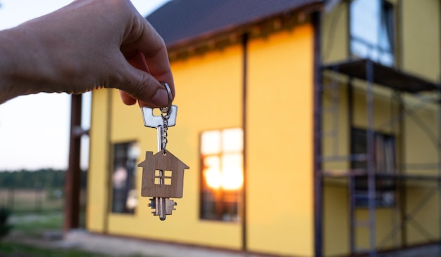 A hand with the keys to a new house on the background of an\
unfinished cottage building project moving to a new home mortgage\
rent and purchase real estate to open the door copy space