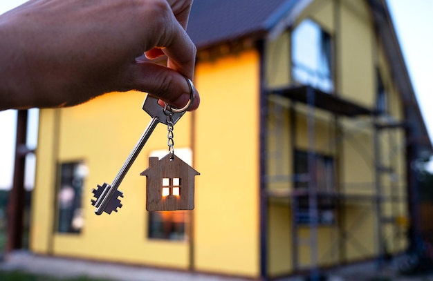 A hand with the keys to a new house on the background of an\
unfinished cottage building project moving to a new home mortgage\
rent and purchase real estate to open the door copy space