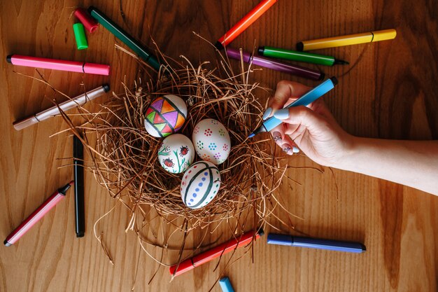 Foto mano con un pennarello sullo sfondo di uova di pasqua colorate che giacciono in un nido su uno sfondo di legno. i marcatori sono sparsi ovunque