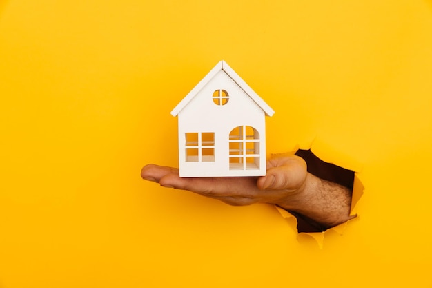 Hand with mini house through a hole in yellow paper background