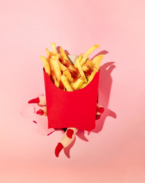 Hand with red nails holding french fries in red paper box pink background Halloween surreal idea