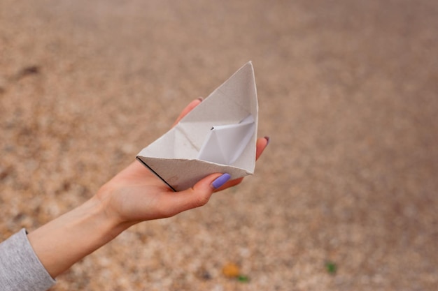 Hand with small paper boat from paper on sand near sea waves\
concept paper boat on a sandy beach near the sea on autumn or sunny\
day travel tourism vacation rest holilday