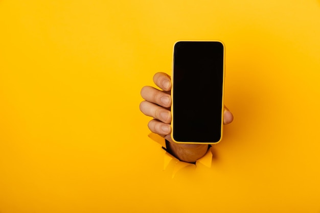 Photo hand with a smartphone out of a hole torn in yellow paper wall