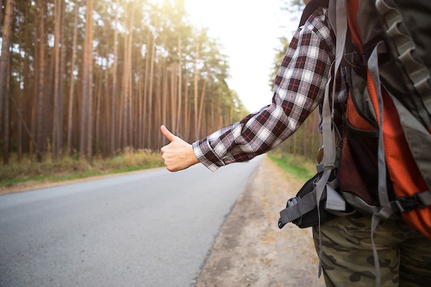 Photo hand with thumbs up on the background of highway - hitchhiking, voting. tourist in a check shirt with backpack near the forest hitch a ride. hitchhiking, domestic tourism, adventure alone, trip, hike