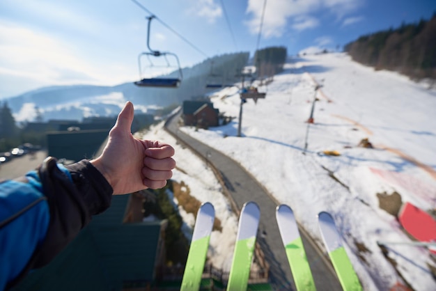 Hand with thumbup gesture and skis on background of mountain skilift rope way on winter day