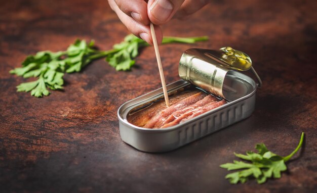 Photo hand with toothpick pricking canned anchovies on a brown table with parsley