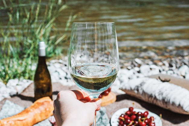 hand with white wine glass on the river coast and picnic background