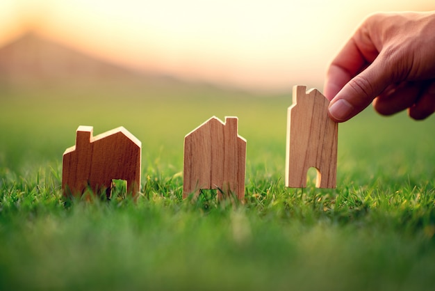 Рука женщины выбирая мини модель деревянного дома на зеленой траве