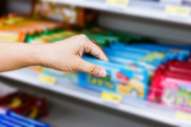 hand of woman choosing or taking sweet products, snacks on shelves in convenience store