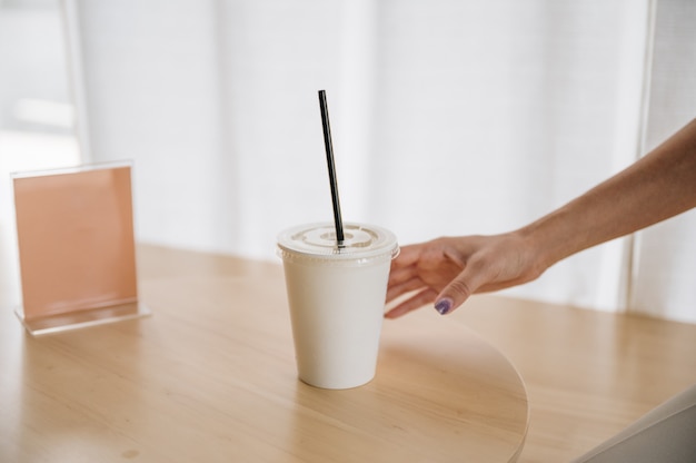 Рука женщины, держащей одноразовую кофейную чашку на деревянном столе с пустой этикеткой в кафе