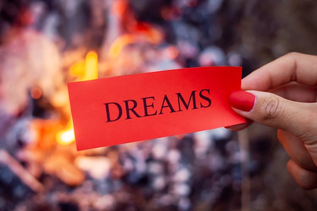 Hand of a woman holding red small sheet of paper with word "dreams" on it with bonfire on background. Symbol of burning dreams, that didn't come true.