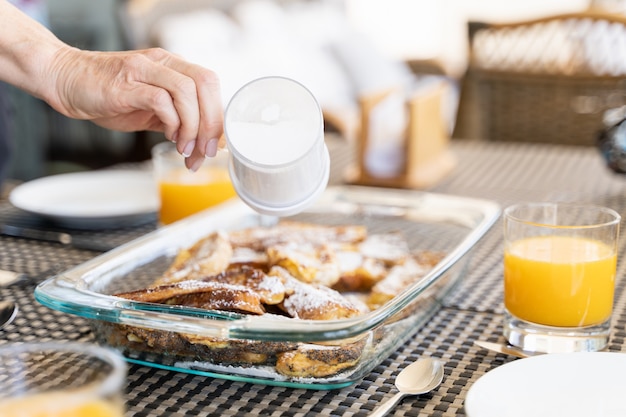 Hand of woman pouring powdered sugar on french toasts glass tray by orange juices on breakfast table