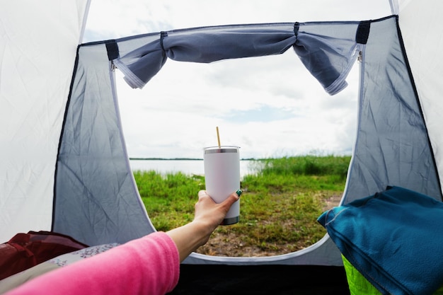 The hand of a woman with a thermocup who is sitting in a tent with a beautiful view of the lake