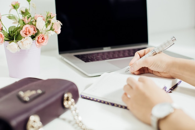 노트북, 꽃 냄비가 있는 사무실 데스크탑에 빈 메모장에 글을 쓰는 여성의 손