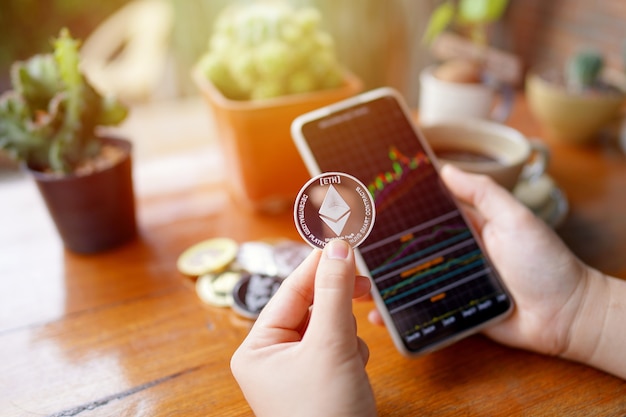 Hand of womans holding ethereum coin and smartphone showing stock chart in cafe